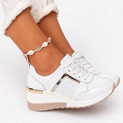 A white women's wedge trainer with laces, perforations and gold metal trim